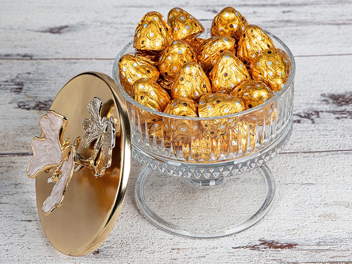 White washed wood with crystal glass jar with gold foil wrapped chocolate and elegant gold lid leaning on the side.